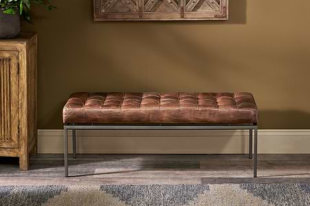 The Nasan Leather Upholstered Bench by nkuku, featuring tan leather and a solid iron frame, is placed against a beige wall with a wood-patterned floor, next to a wooden cabinet. A decorative wall piece hangs above and part of a patterned rug appears in front.