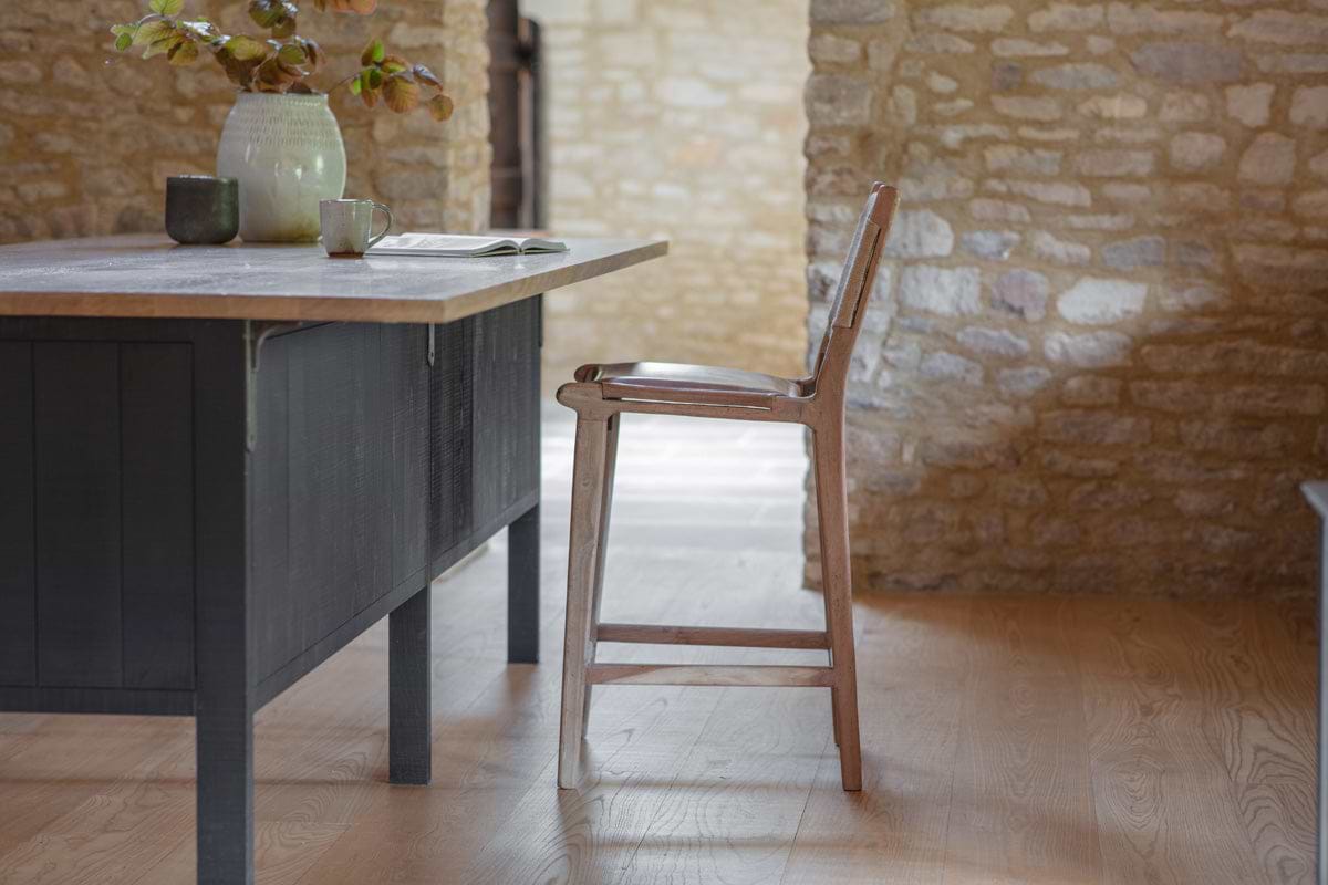 The Nayan Leather & Jute Counter Chair - Tan by nkuku sits by a black kitchen island with a light wood countertop, featuring a ceramic vase with green branches, a cup, and an open book. Stone walls glow in natural light on wood floors.