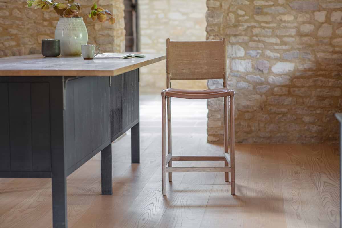 The nkuku Nayan Leather & Jute Counter Chair in tan stands by a large table with a dark base and light wood top, which holds ceramic vases, a mug, and an open book. Stone walls and light wood floors complete the rustic setting.