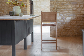 The Nayan Leather & Jute Counter Chair by nkuku sits at a wooden table in a rustic room with light wood floors and exposed stone walls, styled with a ceramic pitcher of branches, two cups, and an open book in soft natural light.