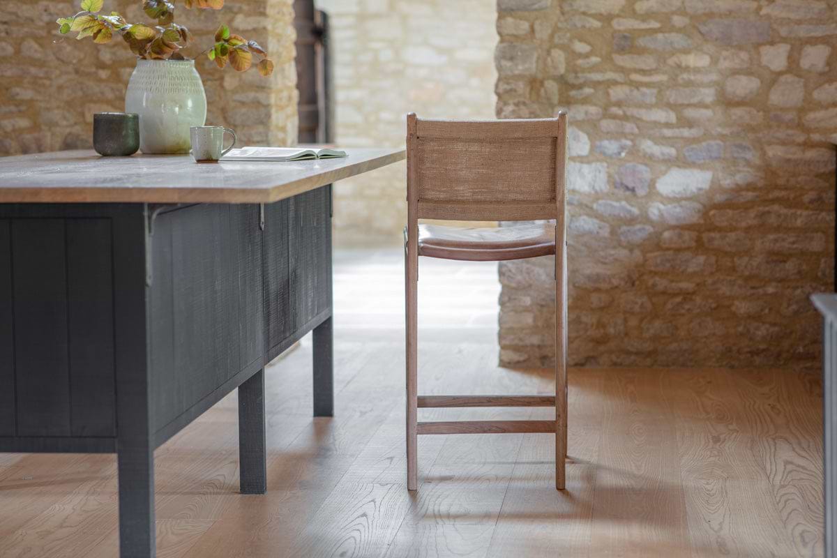 The Nayan Leather & Jute Counter Chair by nkuku sits at a wooden table in a rustic room with light wood floors and exposed stone walls, styled with a ceramic pitcher of branches, two cups, and an open book in soft natural light.