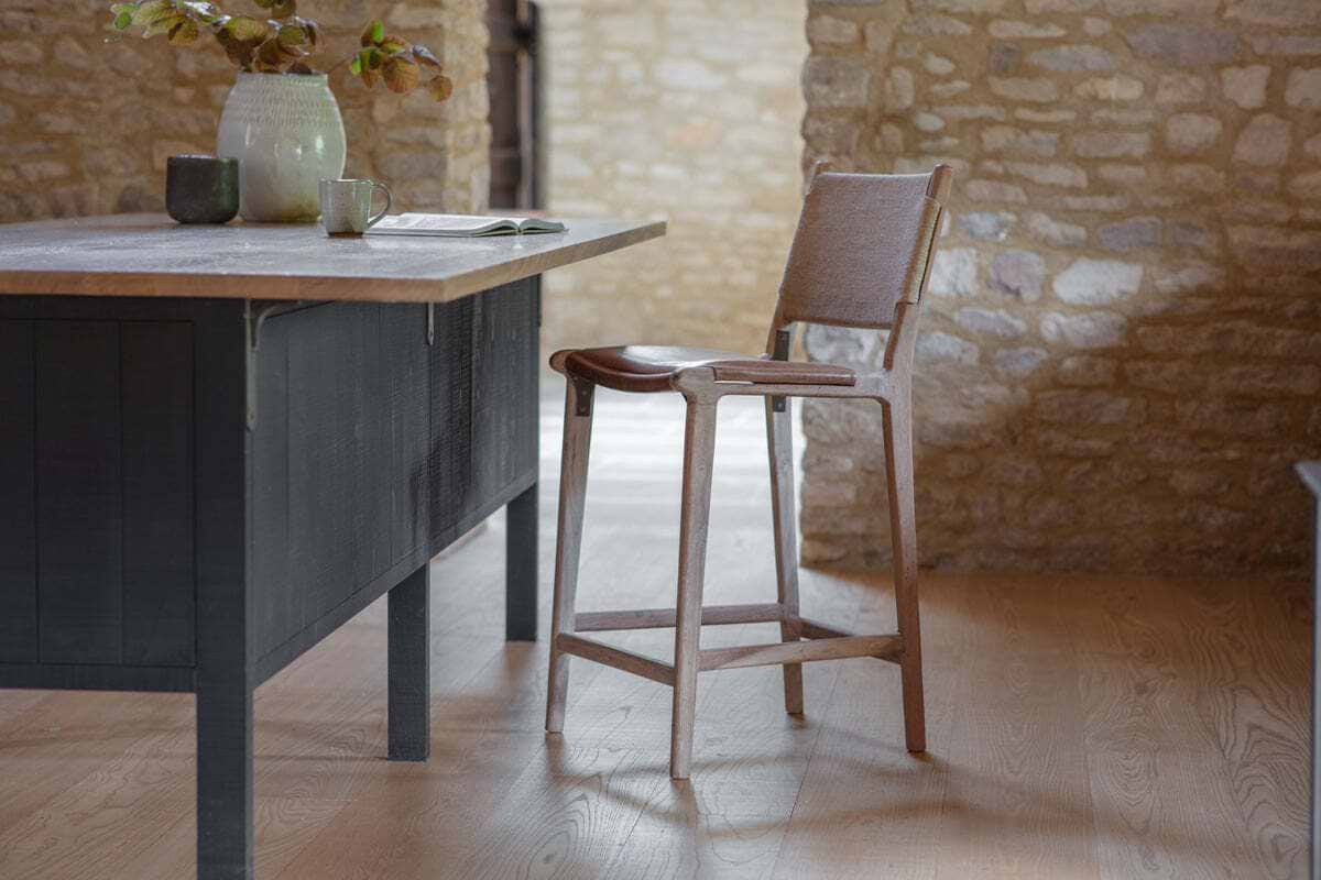 A rustic kitchen features the nkuku Nayan Leather & Jute Counter Chair - Tan beside a dark island with a light stone countertop, ceramic vase of leafy branches, mug, and open book. Stone walls and light wood floors add to the cozy atmosphere.