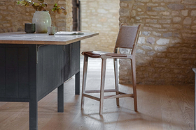 A rustic kitchen features the nkuku Nayan Leather & Jute Counter Chair - Tan beside a dark island with a light stone countertop, ceramic vase of leafy branches, mug, and open book. Stone walls and light wood floors add to the cozy atmosphere.