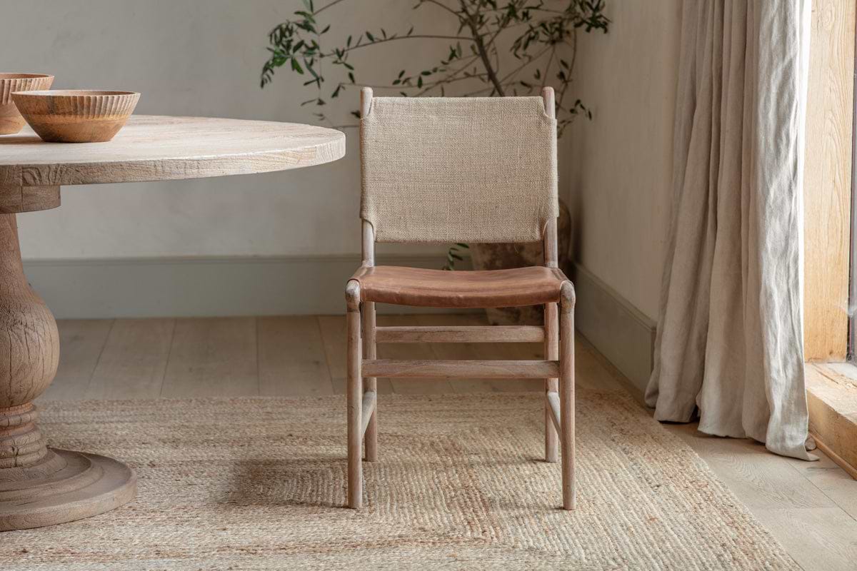 The Nayan Leather & Jute Dining Chair in Tan by nkuku stands on a textured rug beside a round wooden table. Sunlight filters through sheer curtains, while a potted plant and wooden bowls lend natural, minimalist style to the room.