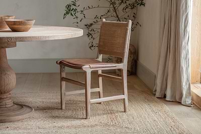 The Nayan Leather & Jute Dining Chair in tan by nkuku sits beside a round wooden table. Light walls, a woven rug, a corner plant, and sheer curtains complete the space’s cozy, natural vibe.