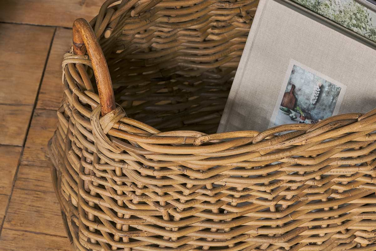 A close-up of the nkuku Netra Rattan Basket - Natural on a wooden floor. This decorative organiser features an interlaced textured pattern, sturdy handle, and holds a fabric-covered book with a vase and dried flowers on its cover.