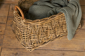 The Netra Rattan Basket - Natural by nkuku sits on a wooden floor, its sturdy handle and chunky-knit greenish-grey blanket draped over the edge bringing a cozy, homey feel to any space.