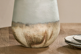 A close-up of the nkuku Ngolo Recycled Glass Vase in Aged Smoke sits on a wooden table beside stacked, white-edged ceramic plates and a spoon. The vase features an earthy, matte finish that blends from white at the top to brown below.