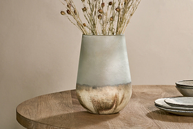 The Ngolo Recycled Glass Vase in Aged Smoke by nkuku sits on a wooden table, holding dried grasses and seed pods. Nearby, two stacked ceramic plates rest against a neutral beige wall.