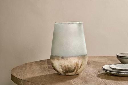 The Ngolo Recycled Glass Vase - Aged Smoke by nkuku, with its gold metallic base, sits on a round wooden table. Stacked ceramic plates with dark rims are placed to the right, set against a plain beige background for a minimalist look.