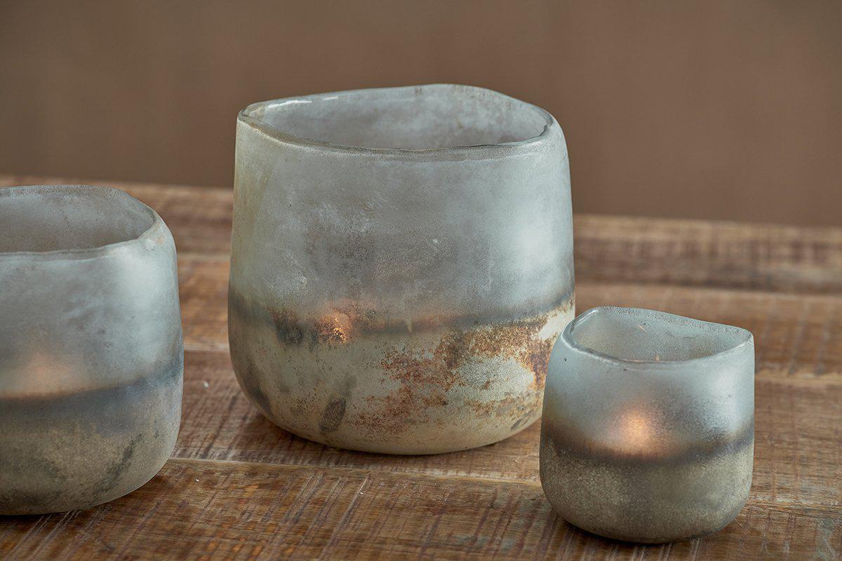 Three Ngolo Tealight Holders by nkuku, crafted from recycled glass in aged smoke tones, sit on a rustic surface. Their handmade look and muted gray hues give off atmospheric light, especially glowing softly inside the smallest holder.