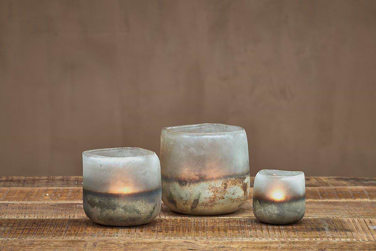 Three uneven Ngolo Tealight Holders in aged smoke from nkuku, crafted from frosted recycled glass, glow with lit candles on a rustic wood surface. The plain brown wall behind enhances their cozy, minimalist atmosphere.