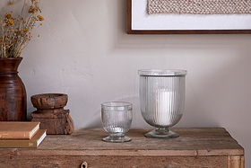 A rustic wooden table displays two nkuku Nia Recycled Glass Candle Holders in clear—one large with a white candle, one small empty. Stacked books, a wooden vessel, dried flowers in a brown vase, and framed textile art sit to the left.