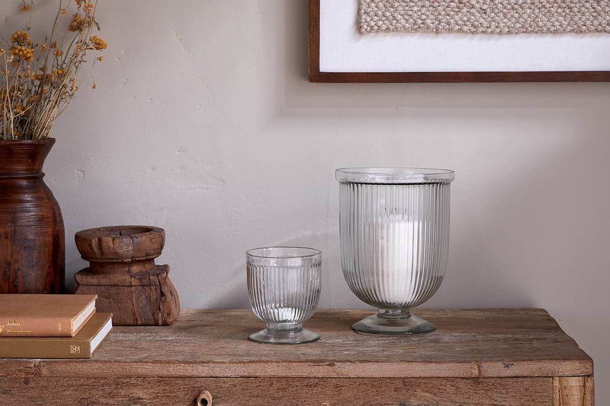 A rustic wooden table displays two nkuku Nia Recycled Glass Candle Holders in clear—one large with a white candle, one small empty. Stacked books, a wooden vessel, dried flowers in a brown vase, and framed textile art sit to the left.
