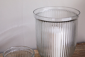 A close-up of nkuku's Nia Recycled Glass Candle Holders in clear, featuring a fluted design on wood—a larger holder with a white candle and the upper edge of the smaller empty holder visible against a plain light wall.
