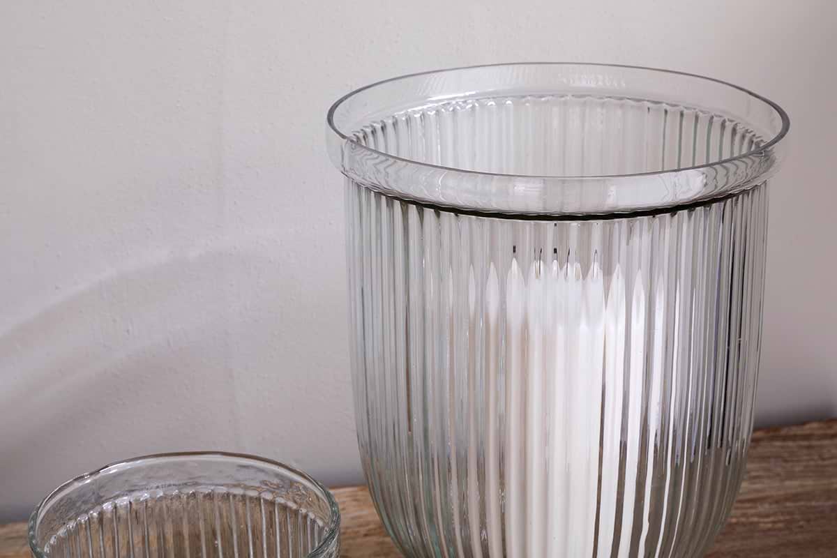 A close-up of nkuku's Nia Recycled Glass Candle Holders in clear, featuring a fluted design on wood—a larger holder with a white candle and the upper edge of the smaller empty holder visible against a plain light wall.