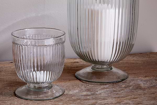 Two Nia Recycled Glass Candle Holders by nkuku, featuring a fluted design and flared bases, sit on a rustic wooden surface. The taller clear holder contains white powder; the smaller is empty. Both can also be used as unique goblets.
