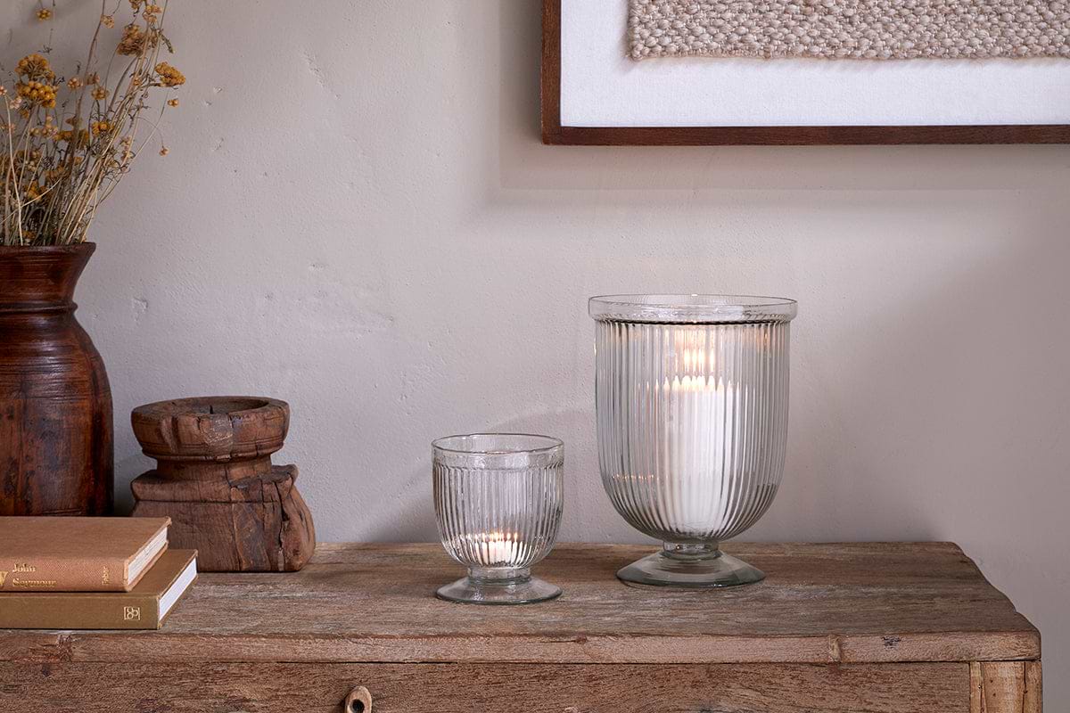 A rustic wooden table displays two nkuku Nia Recycled Glass Candle Holders in clear—one large, one small, both glowing. Nearby are stacked books, a wooden jar, dried yellow flowers in a brown vase, and a textured artwork above.