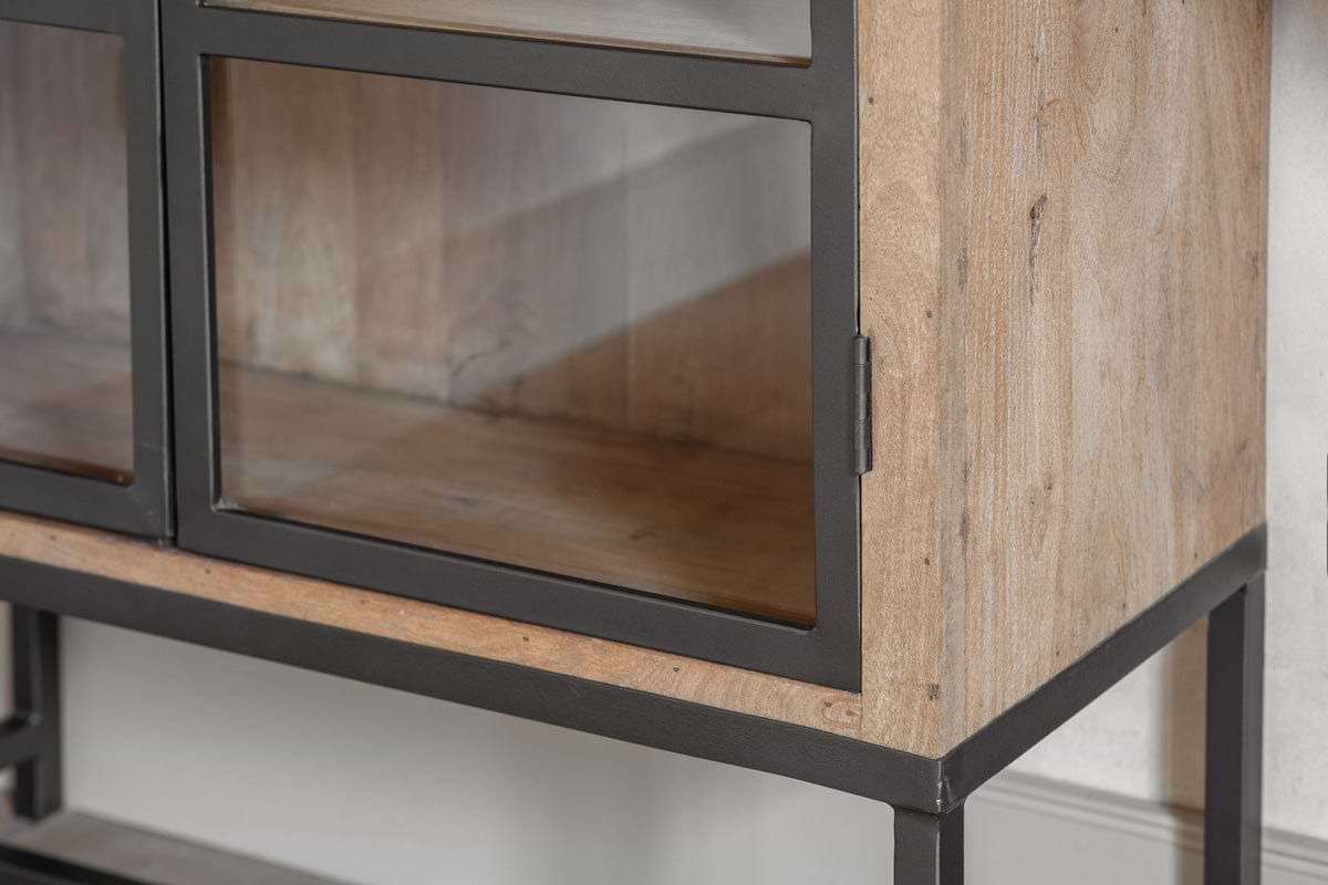 Close-up of the Nirvi Wood & Iron Cabinet - Black by nkuku, featuring mango wood and glass doors with a black metal frame and base. The cabinet showcases industrial style and natural grain details, standing open and empty against a light wall.
