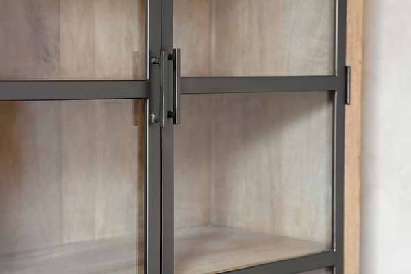Close-up of the nkuku Nirvi Wood & Iron Cabinet - Black, showcasing its antique iron door with a grid over glass and light mango wood shelves inside. The industrial style is balanced by warm wood tones against a neutral background.