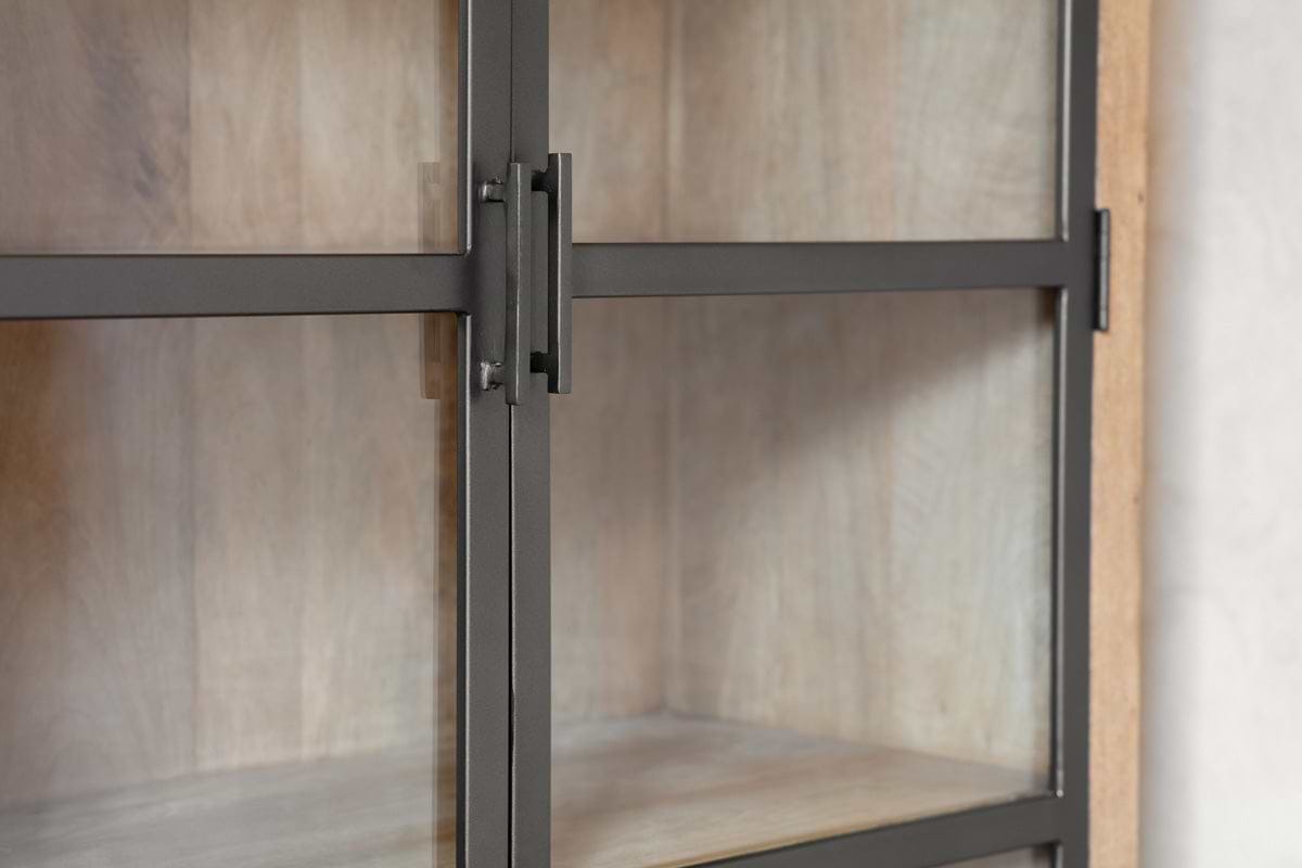 Close-up of the nkuku Nirvi Wood & Iron Cabinet - Black, showcasing its antique iron door with a grid over glass and light mango wood shelves inside. The industrial style is balanced by warm wood tones against a neutral background.