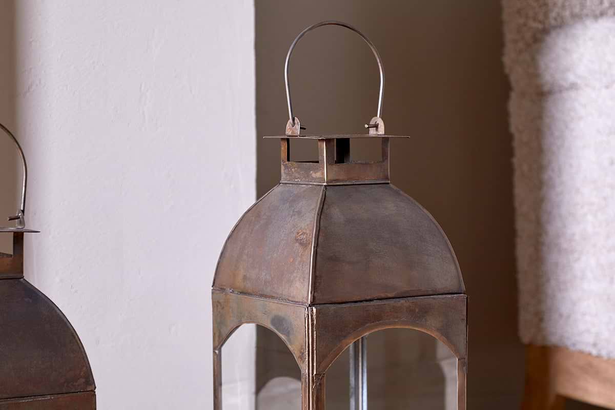 The Nivas Lantern - Antique Brass by nkuku features a rustic metal frame, squared domed top, thin handle, and glass panels with visible patina. It sits against a white wall and beige fabric; part of another lantern appears on the left.