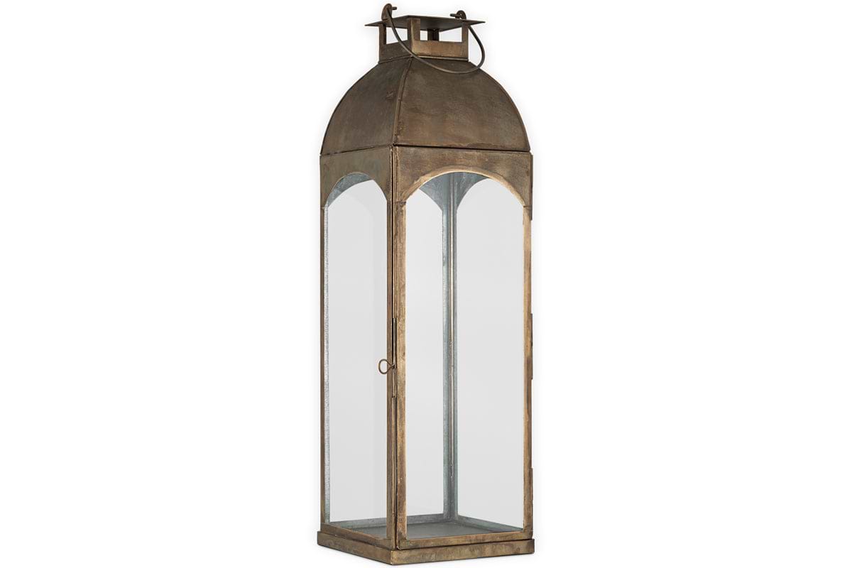 The nkuku Nivas Lantern features an antique brass finish, clear glass panels, arched top, ring handle, rectangular base, and a hinged door for candle placement—perfect as a stylish vintage accent for your home.