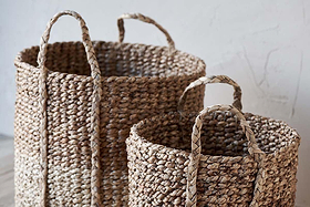 Two nkuku Nkomi Baskets, hand woven from natural textured fibers, sit side by side on a light floor. Both cylindrical storage baskets feature sturdy handles; the larger one is behind the smaller, set against a pale wall.