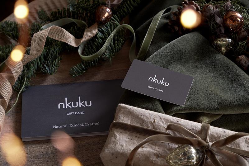 A Nkuku E-Gift Card and envelope sit on wood, surrounded by green ribbon, pine branches, pinecones, and ornaments. Nearby is a string-tied brown-paper gift, creating a festive and rustic holiday mood.