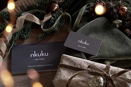 A Nkuku E-Gift Card and envelope sit on wood, surrounded by green ribbon, pine branches, pinecones, and ornaments. Nearby is a string-tied brown-paper gift, creating a festive and rustic holiday mood.