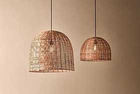 Two Noko Wicker Dome Pendant lights by nkuku hang from the ceiling against a beige wall. The larger pendant is on the left, the smaller on the right—both casting warm, elegant light with exposed bulbs and dome-shaped wicker shades.