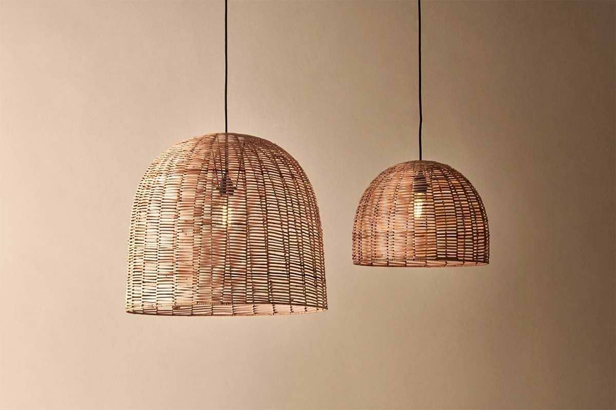 Two Noko Wicker Dome Pendant lights by nkuku hang from the ceiling against a beige wall. The larger pendant is on the left, the smaller on the right—both casting warm, elegant light with exposed bulbs and dome-shaped wicker shades.