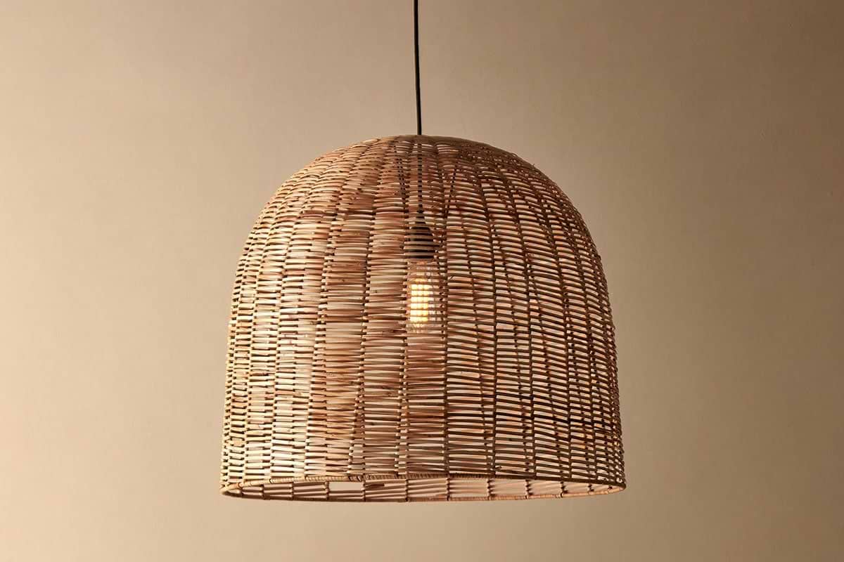 The Noko Wicker Dome Pendant - Large by nkuku hangs from a black cord against a beige wall, featuring an exposed bulb shining through its natural woven shade—a stylish lighting option for any room.