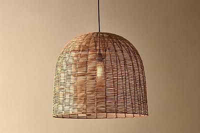 The Noko Wicker Dome Pendant - Large by nkuku hangs from a black cord against a beige wall, featuring an exposed bulb shining through its natural woven shade—a stylish lighting option for any room.