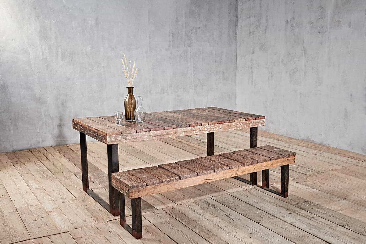 The nkuku Oso Wooden Bench sits beside a rustic wooden table with metal legs. On the table are a brown bottle with dried wheat stems and two clear glasses, set against a plain gray textured wall and wooden floor.