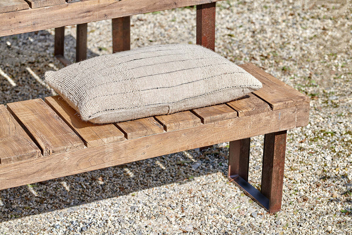 A beige, striped knit cushion sits on the nkuku Oso Wooden Bench with metal legs and a slatted design. Sunlight highlights the textures of wood and fabric as the bench stands outdoors on gravel.