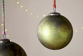 Two large Oko Giant Bauble - Matt Green ornaments by nkuku hang from colorful strings against a neutral background with soft yellow lights. Their slightly tarnished, vintage look adds rustic charm to this festive scene.