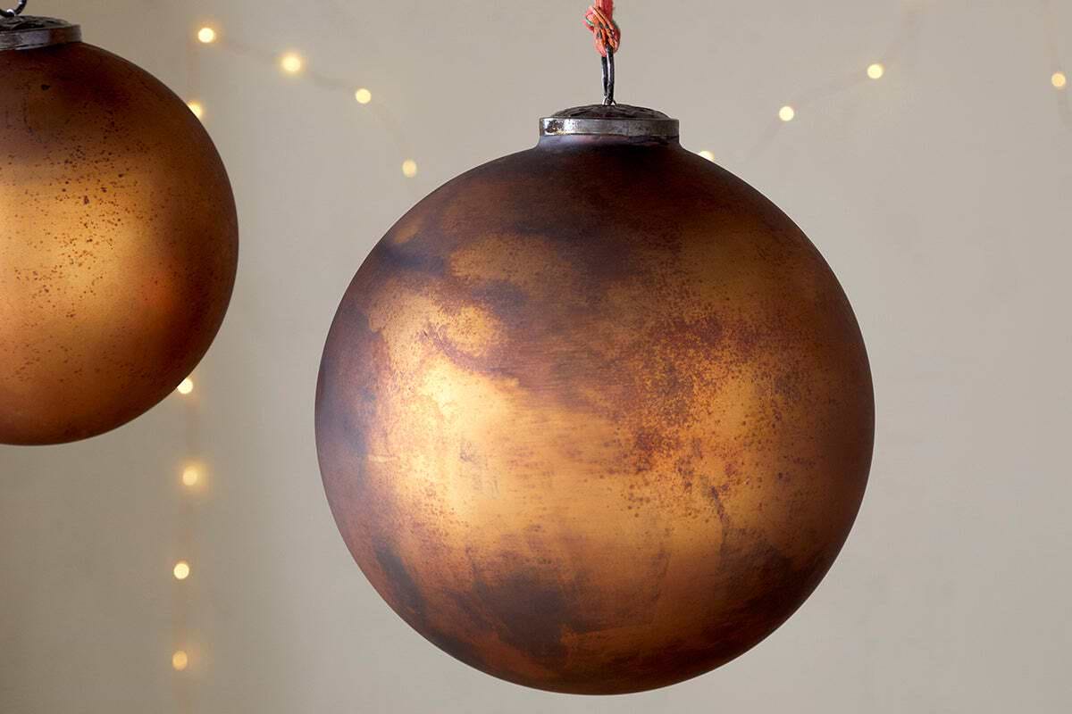A close-up of the nkuku Oko Giant Bauble in matt gold. Warm string lights blur softly in the background, with another similar ornament partially visible among the Christmas decorations.