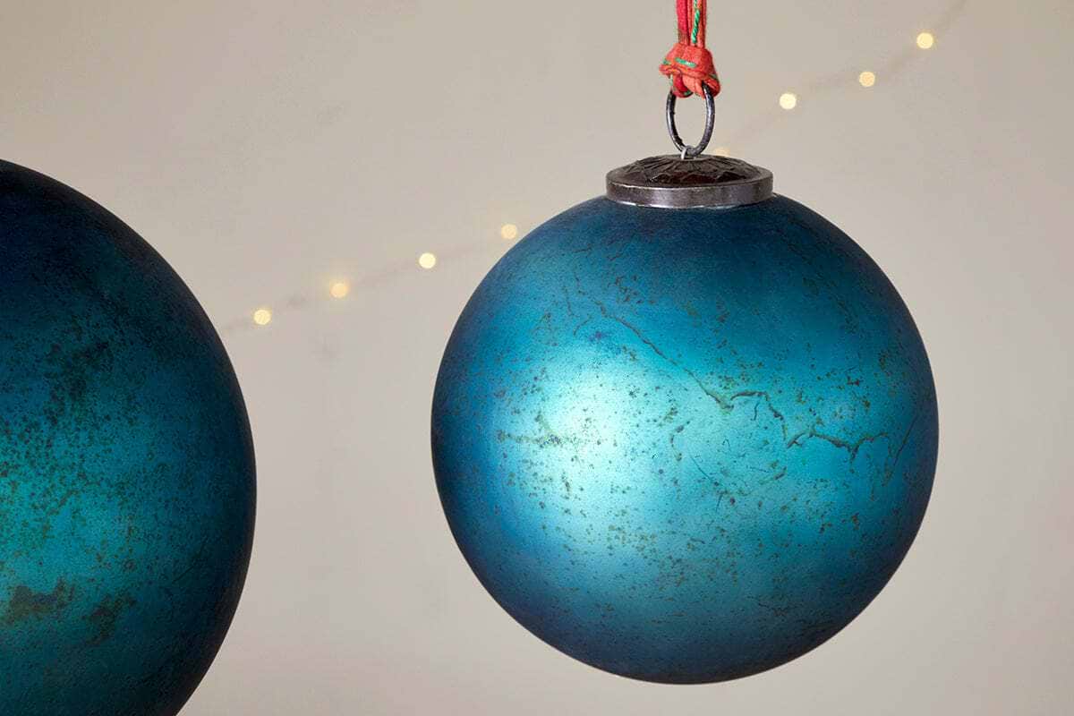 A close-up of two nkuku Oko Giant Bauble - Matt Teal ornaments, one hanging from a red string and the other partially visible, forms a festive scene against a beige background with soft, blurred string lights.