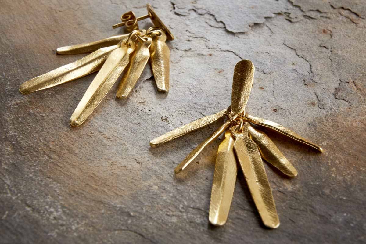The nkuku Omuti Earrings - Gold showcase multiple elongated, textured metal pieces with a gold-coloured finish, styled on a stone surface—ideal for lovers of contemporary jewellery.