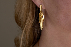 Close-up of a person wearing the Omuti Earrings - Gold by nkuku, featuring multi-layered, leaf-shaped design with a gold finish. The party-ready earrings hang elegantly, with light brown hair and skin in the background.