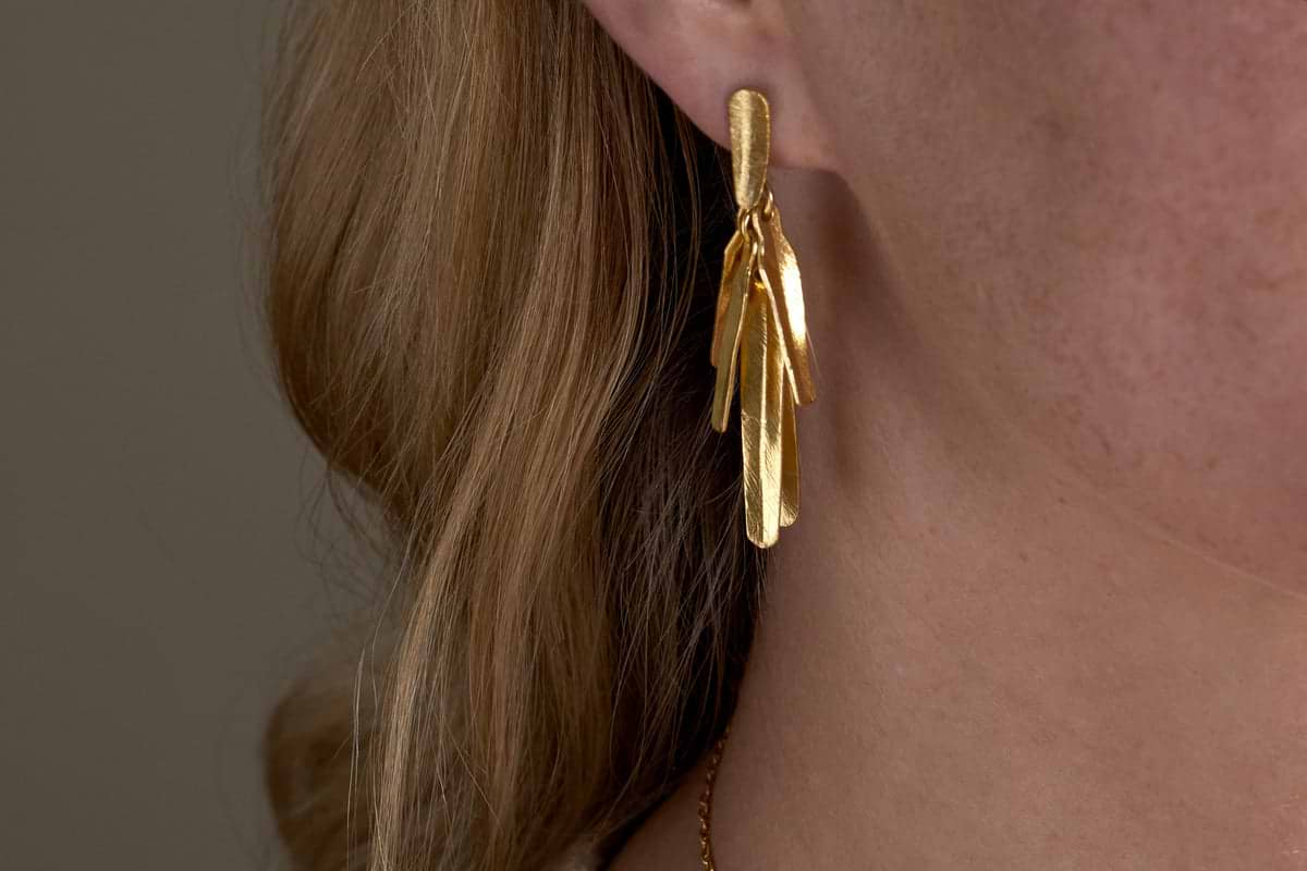 Close-up of a person wearing the Omuti Earrings - Gold by nkuku, featuring multi-layered, leaf-shaped design with a gold finish. The party-ready earrings hang elegantly, with light brown hair and skin in the background.