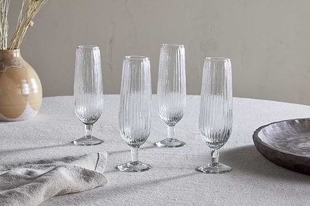 A set of four Otuba Champagne Flutes in clear, by nkuku, are arranged on a light tablecloth, with a ceramic vase of dried plants and a wooden bowl in the background.