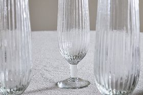 A set of four nkuku Otuba Champagne Flutes in clear glass is arranged on a light grey textured surface, with one flute sharply in focus at the center and the others partially visible on either side.