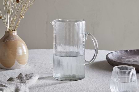An Otuba Jug - Clear by nkuku, half-filled with water, rests on a light-colored textured tablecloth beside a matching glass, a beige ceramic vase with dried flowers, a napkin, and a wooden bowl.