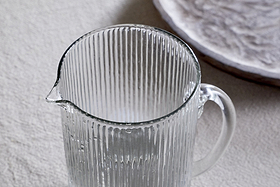 The Otuba Jug - Clear by nkuku, made from recycled glass with a vertical ribbed texture, rests elegantly on a light surface. In the background, a subtly patterned white plate complements its timeless design.