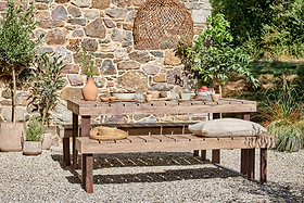 The nkuku Oso Wooden Dining Table with benches stands on gravel by a stone wall, set with ceramic dishes and vases. Bench cushions, green plants, and a woven pendant lamp complete the cozy outdoor scene in natural light.