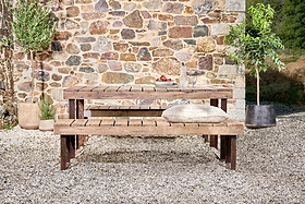 The nkuku Oso Wooden Dining Table and bench set sits on gravel beside a stone wall. On the table are a book, apples, and two glasses. Two potted plants flank the scene, and a beige cushion rests on the bench in soft sunlight.