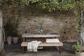 The Oso Wooden Dining Table by nkuku, paired with a matching bench, sits on a brick patio against a weathered wall. A clear vase with leafy branches, dining ware, cushions, and lush plants complete the inviting setup.
