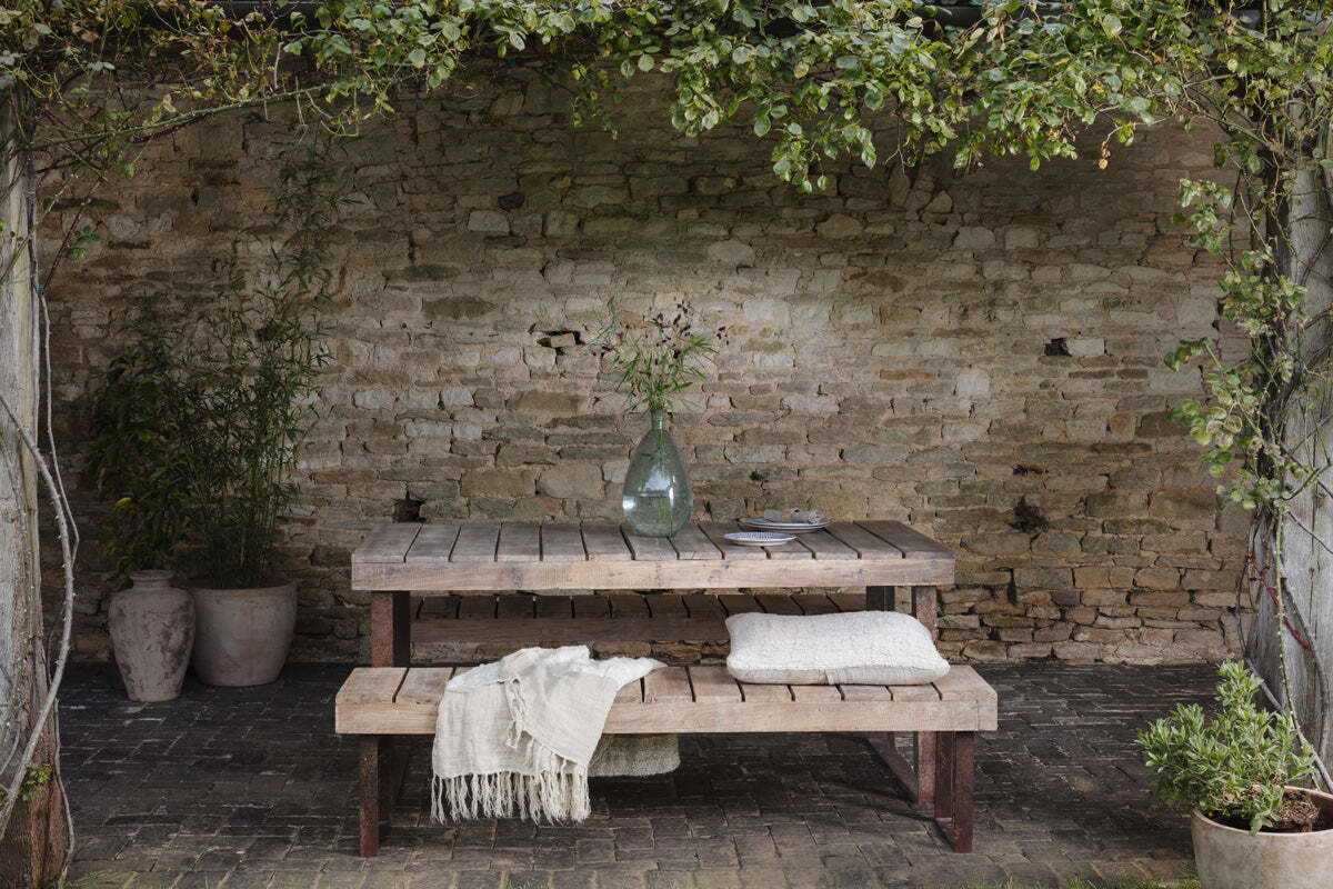 The Oso Wooden Dining Table by nkuku, paired with a matching bench, sits on a brick patio against a weathered wall. A clear vase with leafy branches, dining ware, cushions, and lush plants complete the inviting setup.